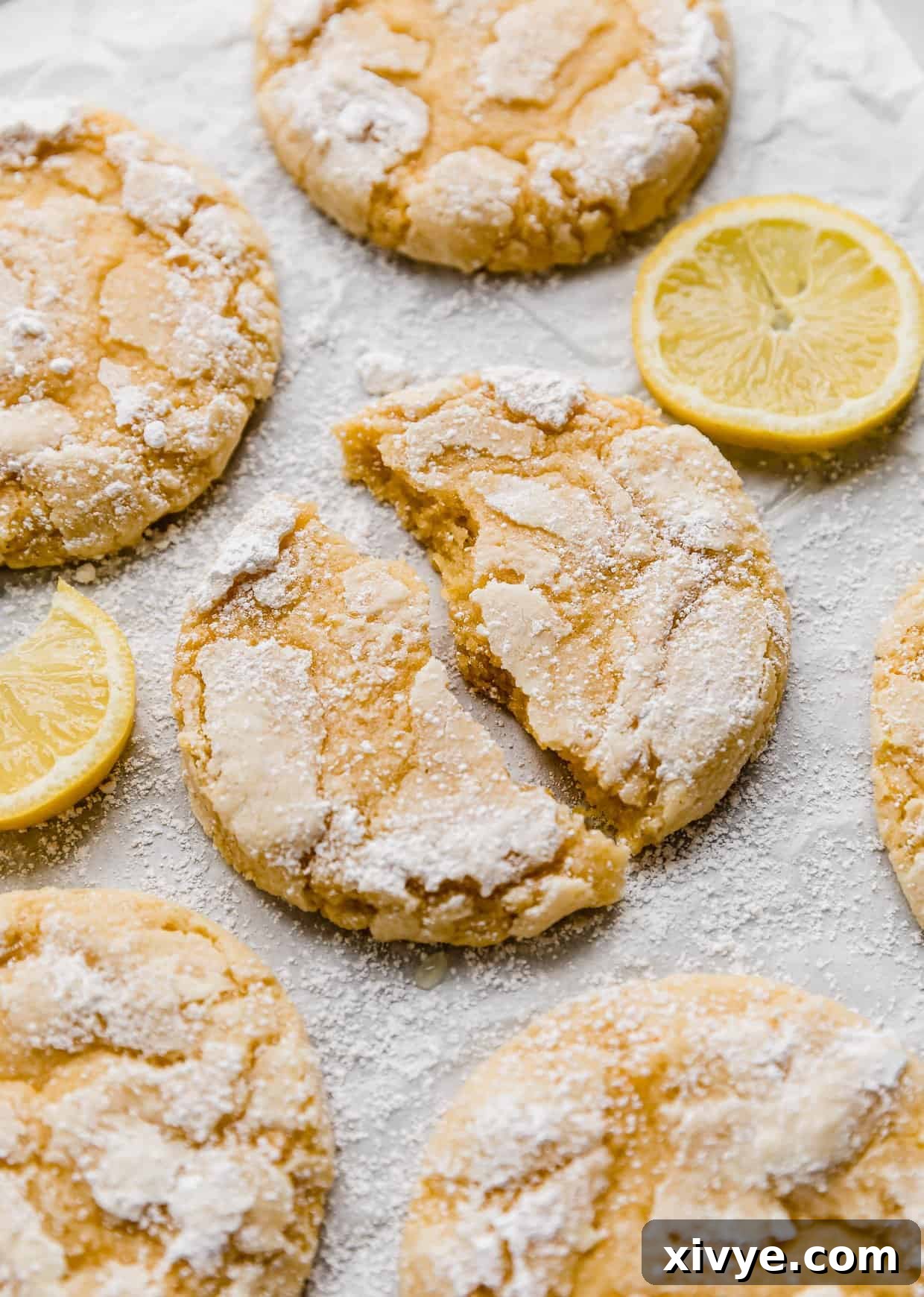 Zesty Lemon Crinkles 11 A Lemon Crinkle Cookie torn in half, on a white background with lemon slices around the cookie, showing its soft interior.