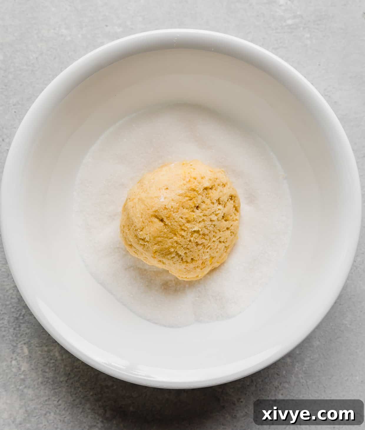 Zesty Lemon Crinkles 6 Lemon Crinkles cookie dough being rolled in a white bowl filled with granulated sugar, showing the initial coating step.