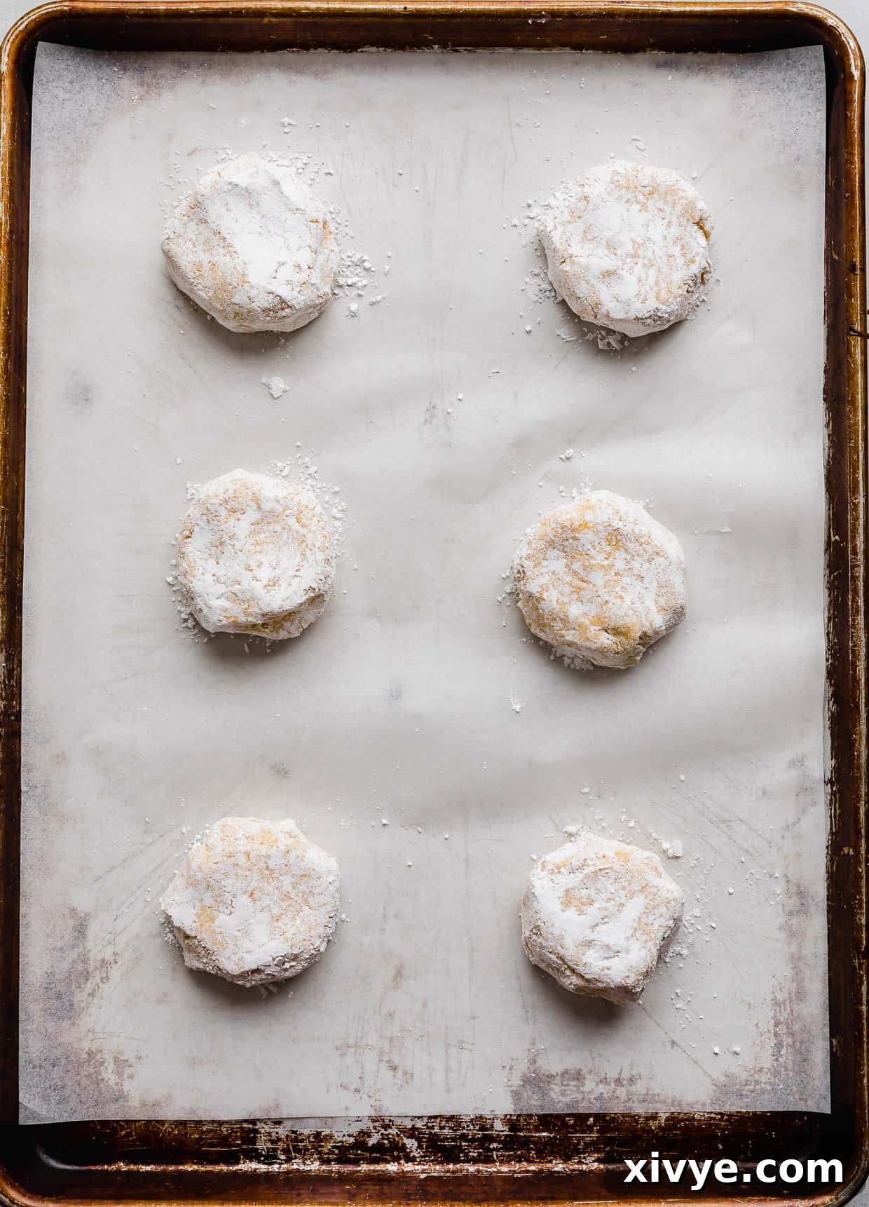Zesty Lemon Crinkles 8 Six lemon crinkle cookie dough balls rolled in powdered sugar on a white parchment lined baking sheet, spaced for baking.