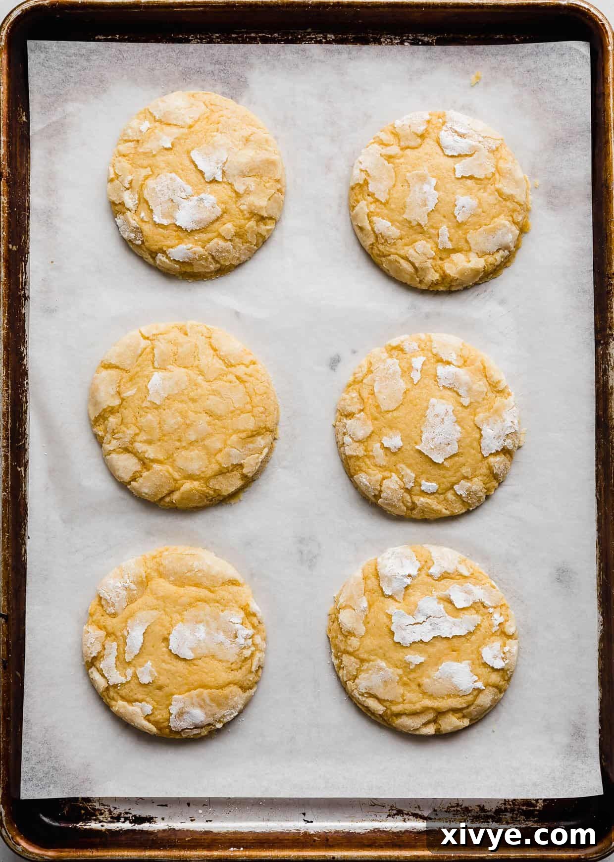 Zesty Lemon Crinkles 9 Six large baked lemon crinkle cookies on a white parchment lined baking sheet, showcasing their crinkled tops.