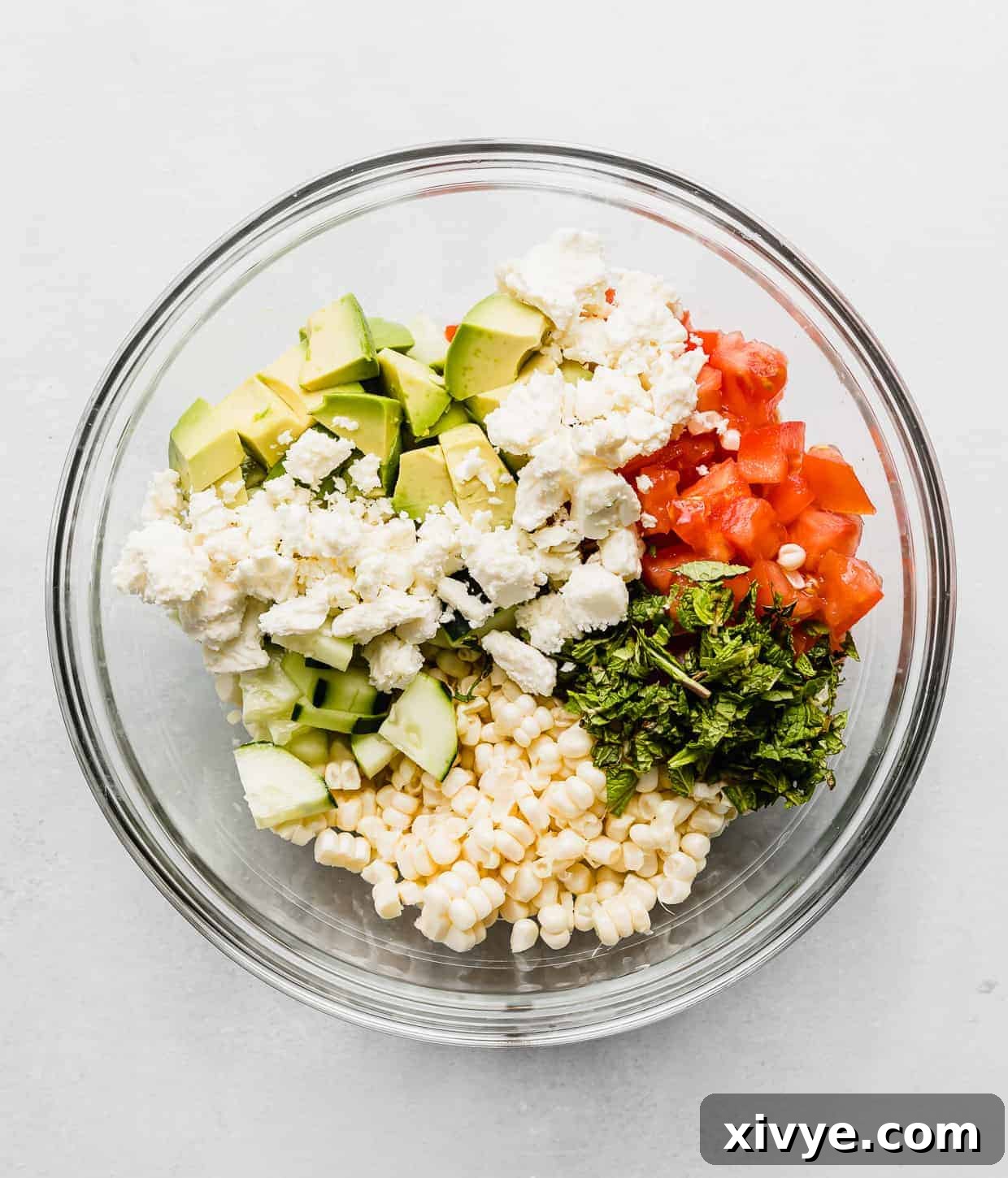 A glass bowl full of corn kernals, diced tomatoes, feta cheese, avocado and mint.
