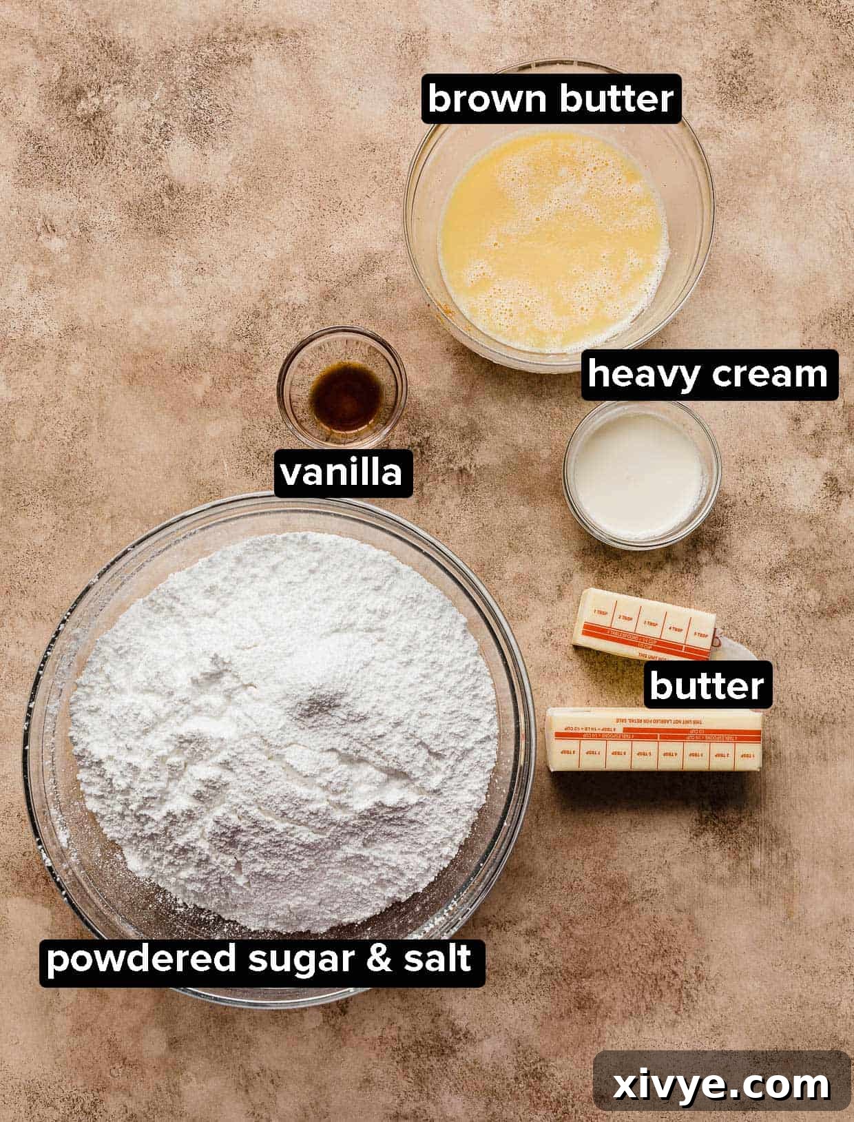 Brown Butter Frosting ingredients on a brown background: powdered sugar, salt, butter, brown butter, heavy cream, and vanilla.