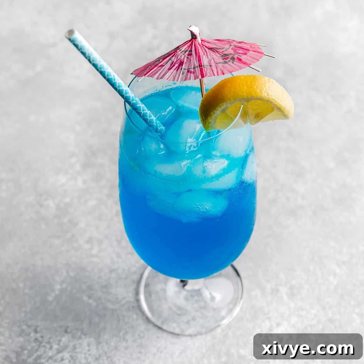 A Blue Lagoon Mocktail in a large glass cup with a pink umbrella and lemon slice on the rim.