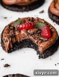 A bite taken out of a chocolate frosting topped Dirt Cake Cookie with a garnish of Oreo crumbs and a gummy worm.