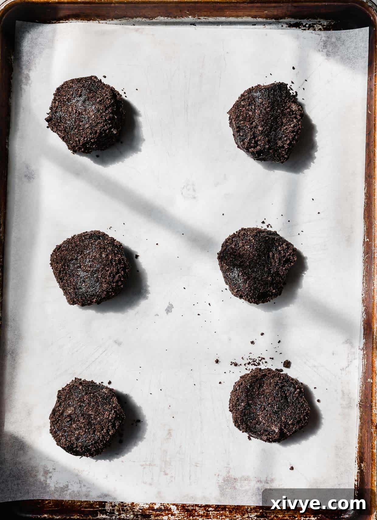 Six black Dirt Cake Cookie dough balls on a white parchment lined baking sheet.
