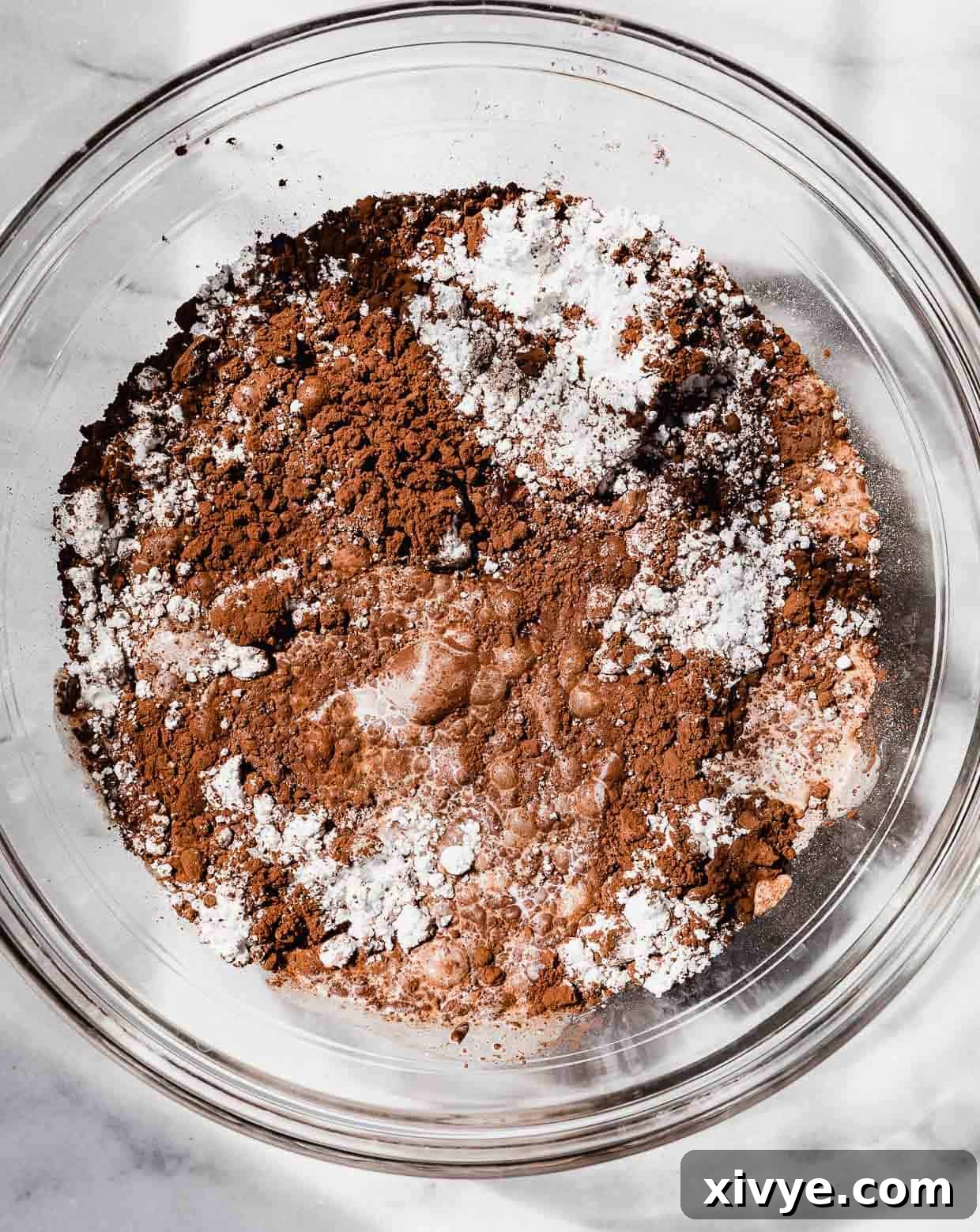 Cocoa powder, powdered sugar, and milk in a glass bowl on a marble background.