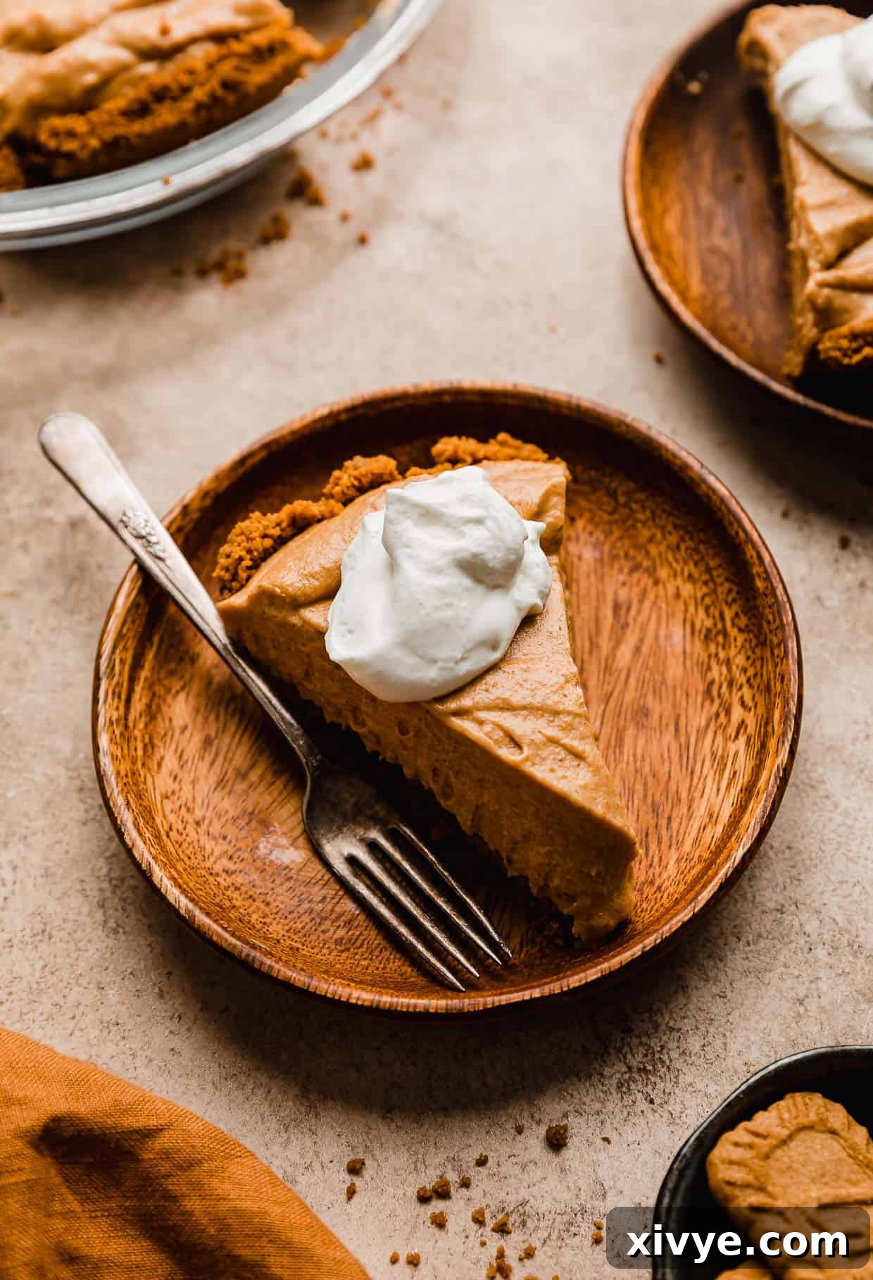 A beautiful slice of Biscoff Pumpkin Pie, showcasing its creamy texture and Biscoff cookie crust, served on a rustic wooden plate.