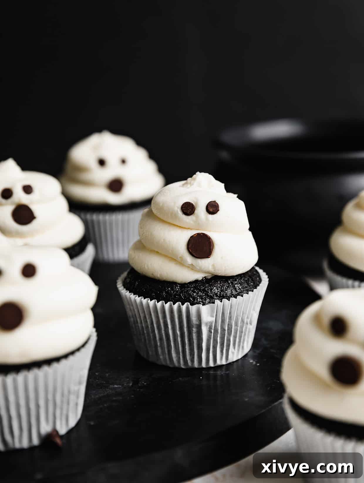 Ghost Cupcakes on a black table against a black background. The white frosting ghosts have chocolate chip eyes and mouths.