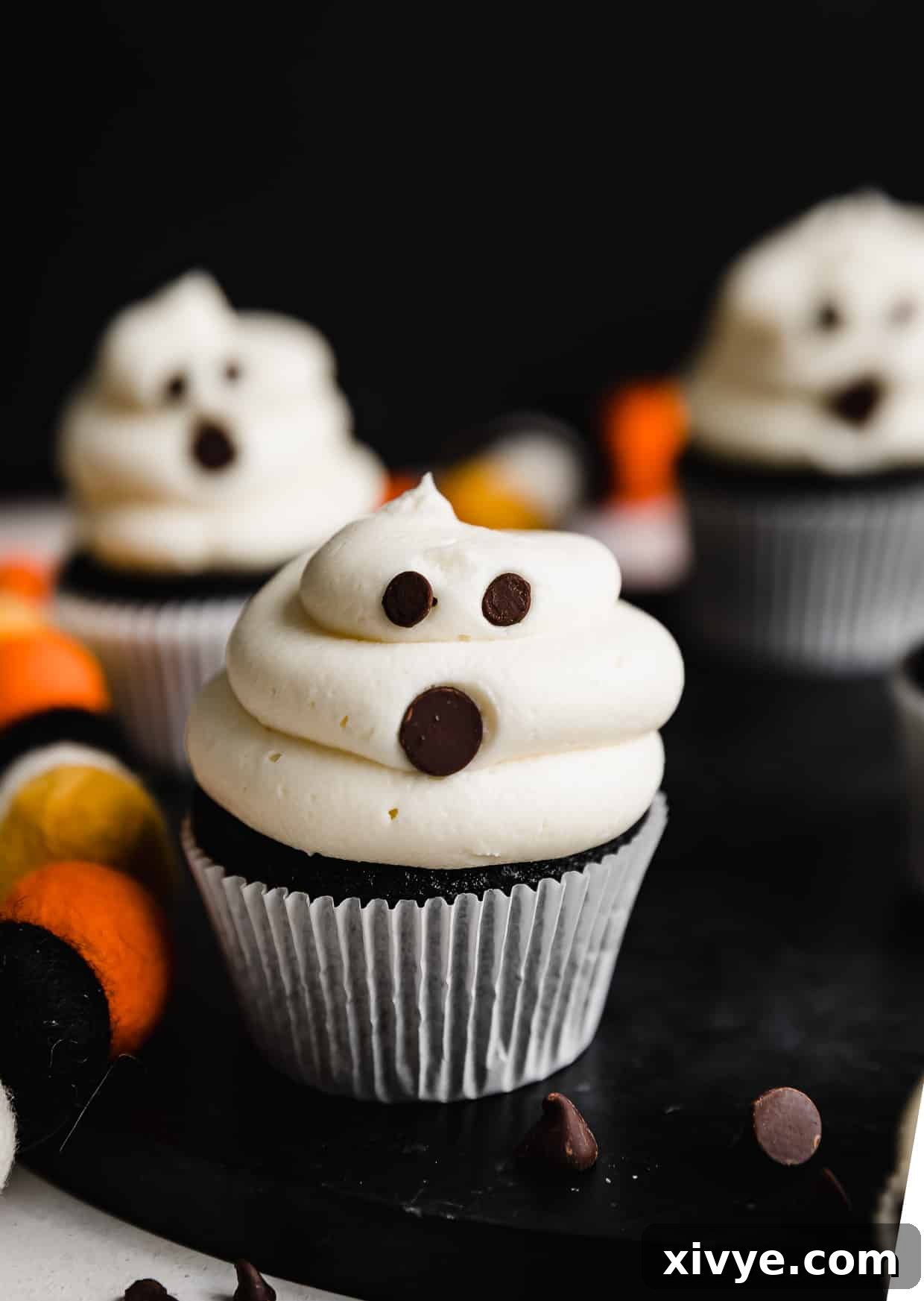White frosting covered cupcakes with chocolate chips stuck into the frosting to make Ghost Cupcakes. The cupcakes are on a black background.