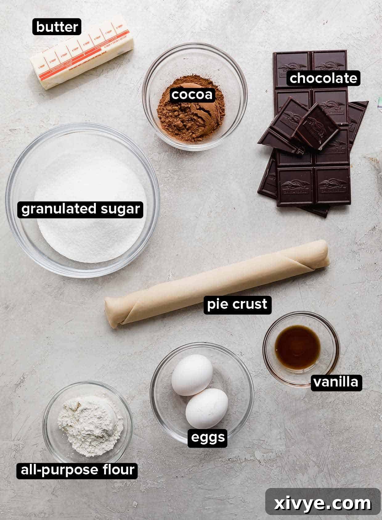 Brownie Pie ingredients on a light gray background, including chocolate, cocoa, pie crust, eggs, vanilla, butter, and sugar.