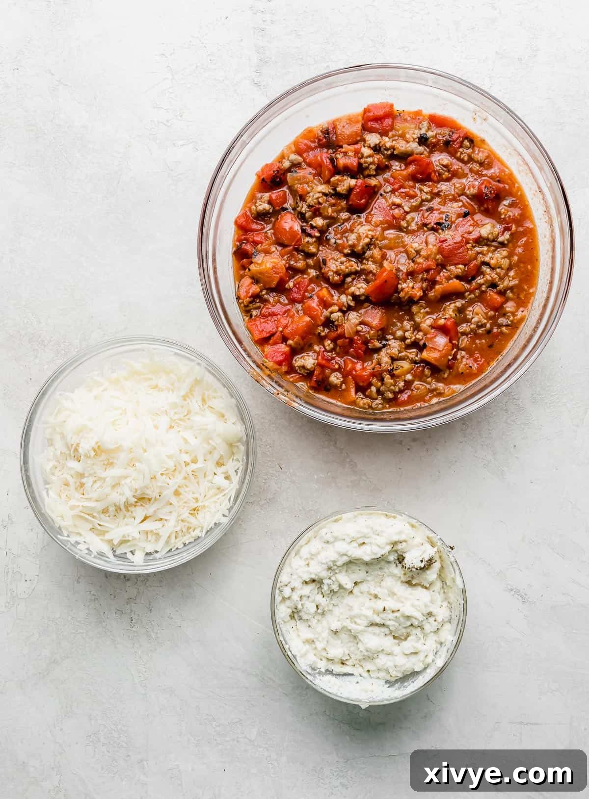 Three bowls: one with red tomato and meat ragu, one with shredded mozzarella and parmesan cheese, and another with an herb ricotta mixture; all are prepped for cast iron lasagna assembly.