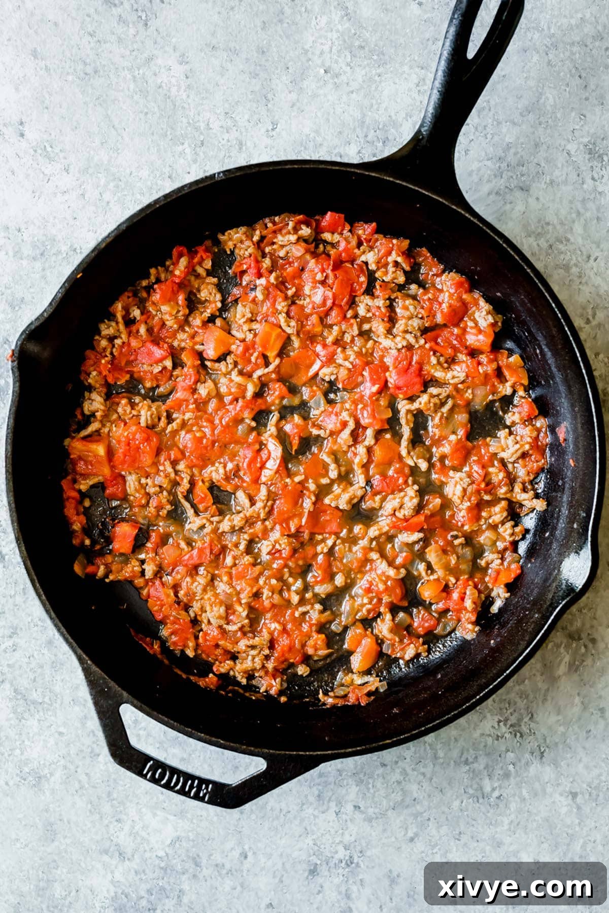 A cast iron skillet with a meat and tomato mixture spread along the bottom, ready for layering.