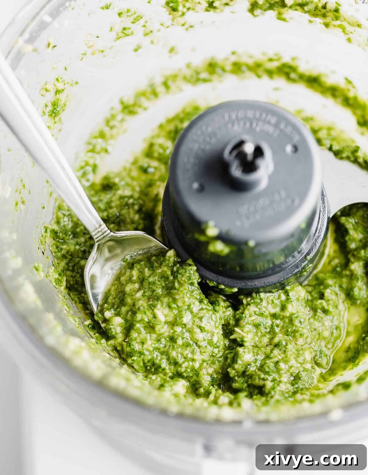 A spoon scooping up fresh basil pesto from a food processor after blending.