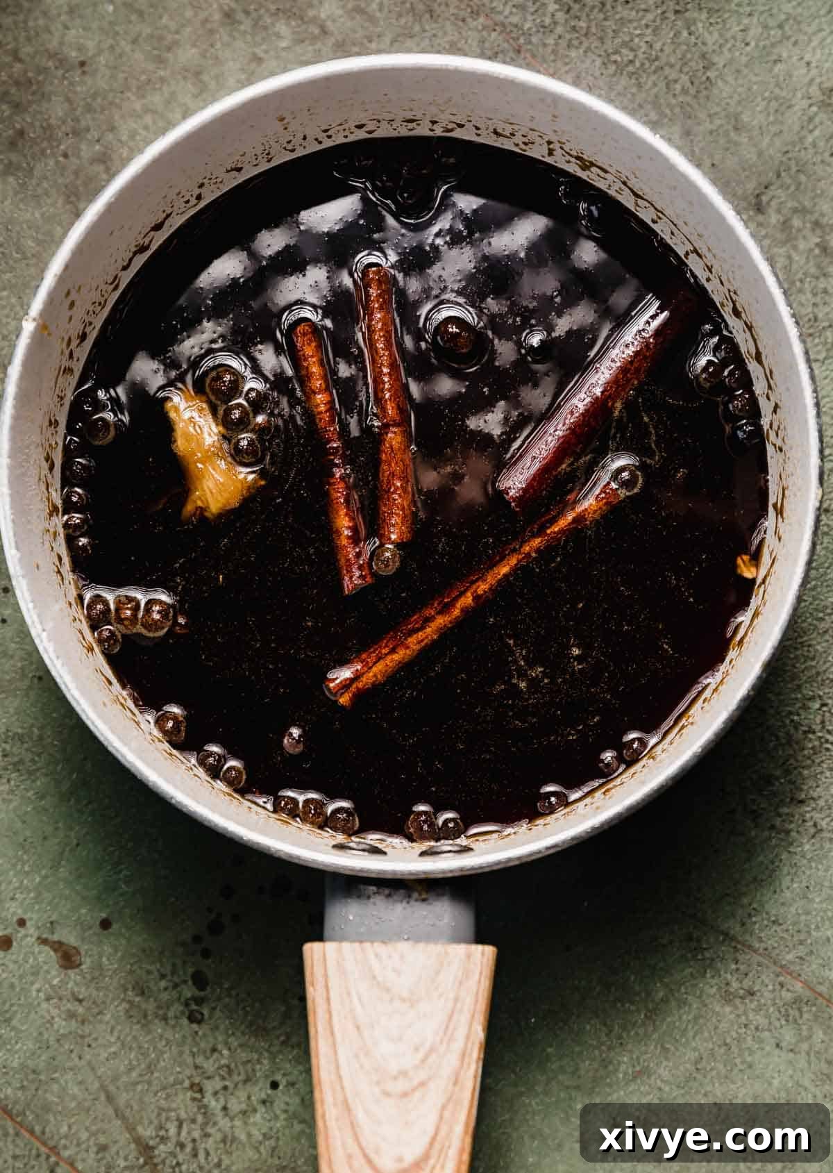 Allspice berries, cinnamon sticks, and fresh ginger in a dark Gingerbread Syrup that's in a saucepan on a green background.