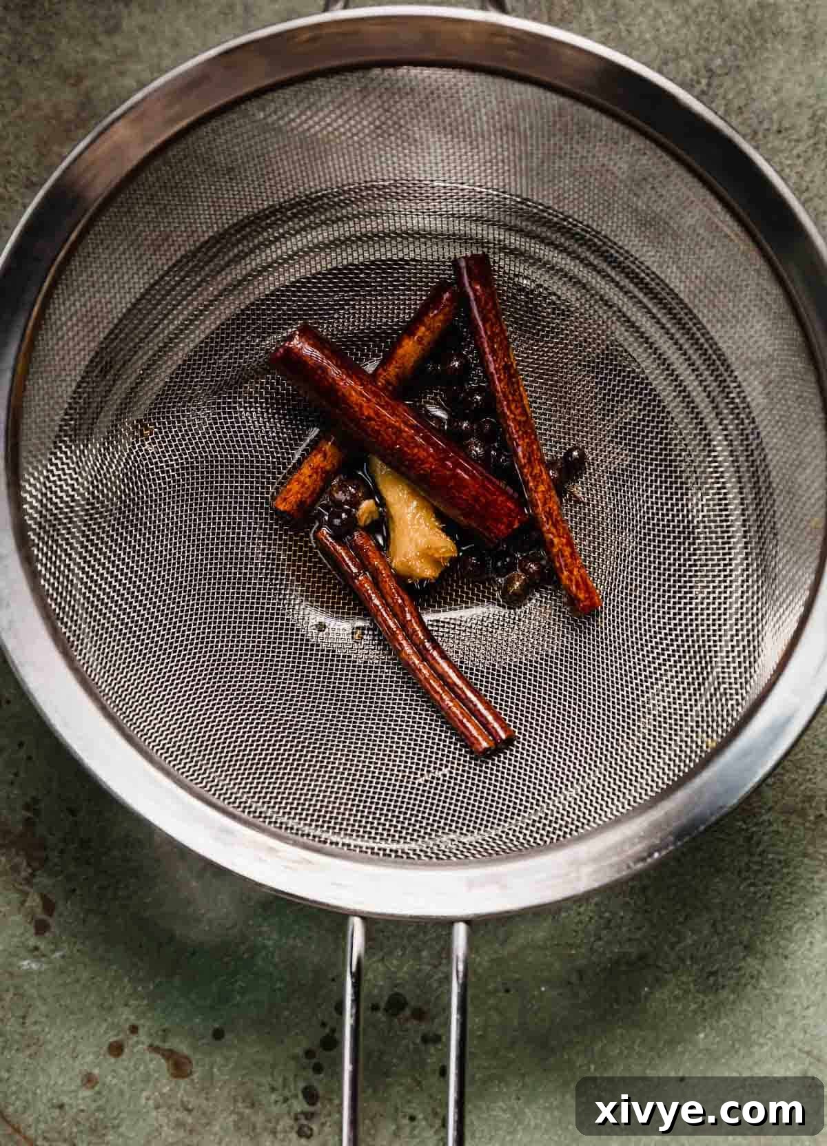 A metal strainer with four cinnamon sticks, handful of allspice berries, and small fresh ginger chunk in it.