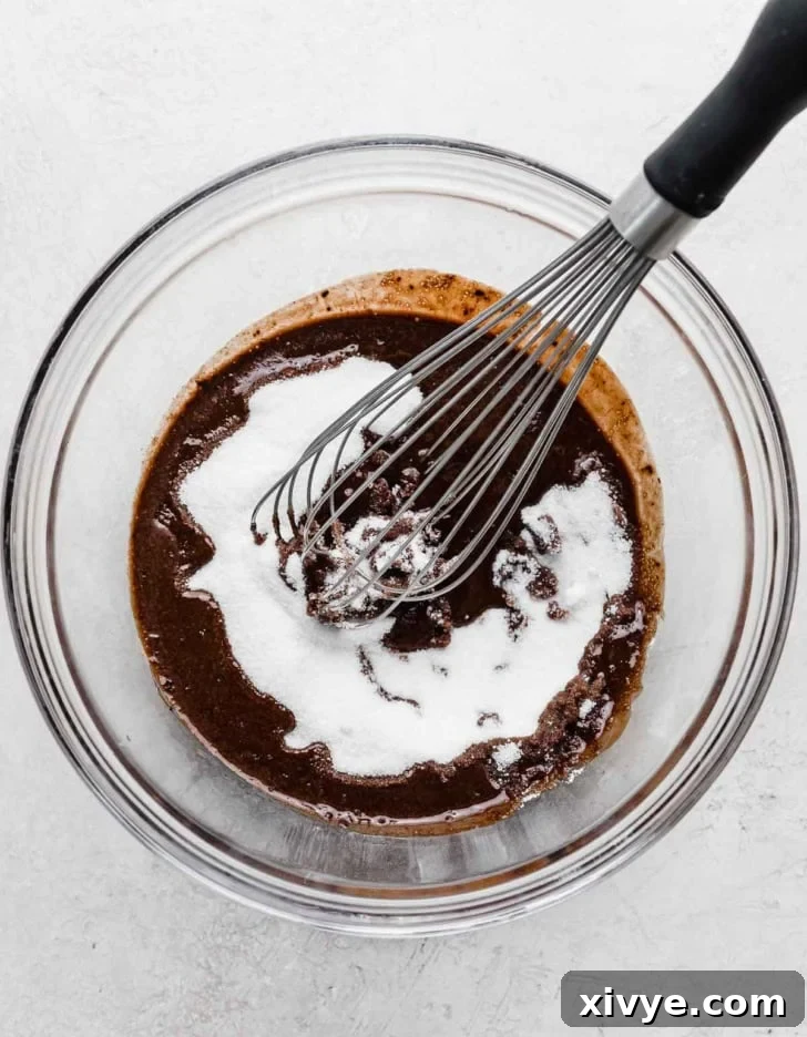 Glass bowl on a gray background with a melted chocolate and white sugar mixture being whisked together.