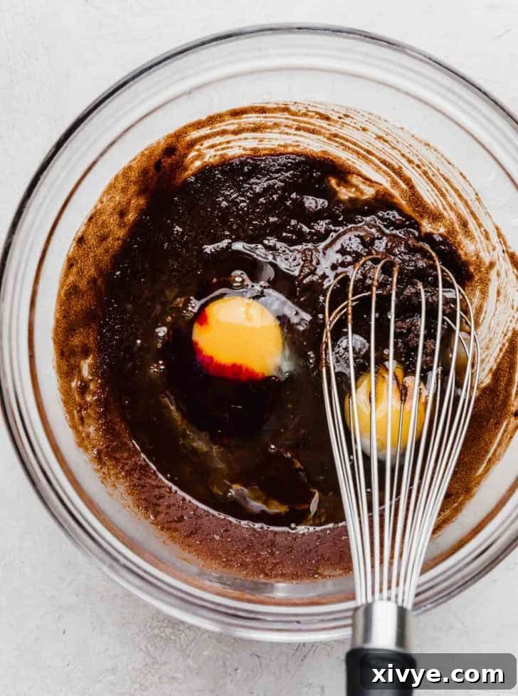 A dark chocolate colored liquid and an egg yolk in a glass bowl with a whisk.