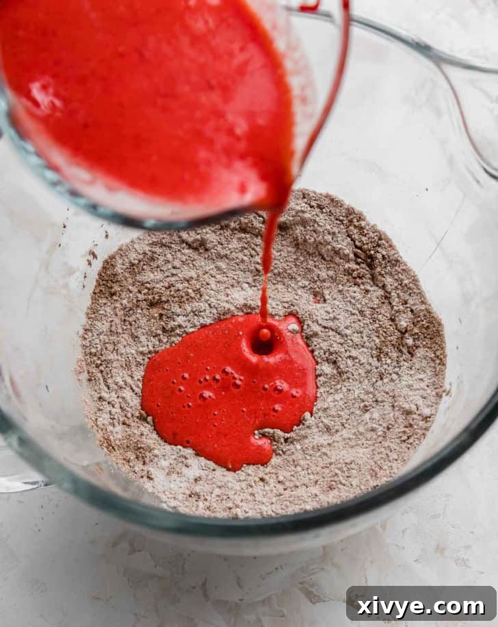 Red liquid being poured into a large bowl filled with red velvet dry ingredients mixture.