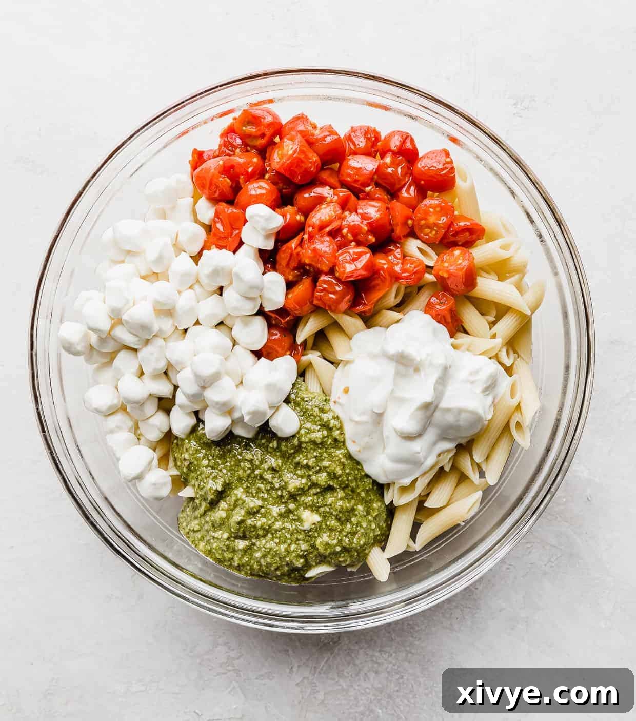 A glass bowl with grape tomatoes, mozzarella balls, sour cream, pesto, a top cooked penne noodles.