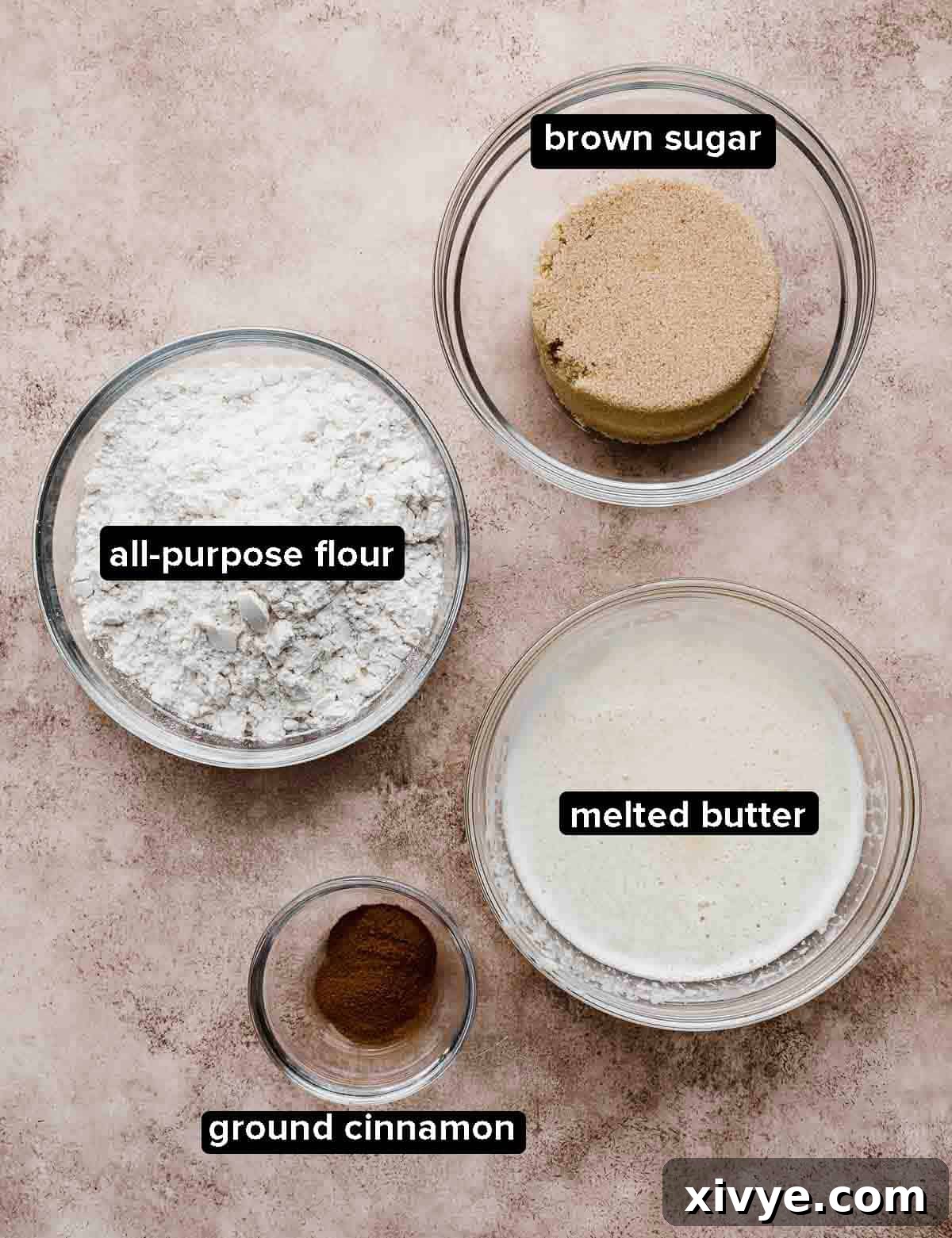 Flour, melted butter, brown sugar, cinnamon in glass bowls on a light brown and beige textured background.