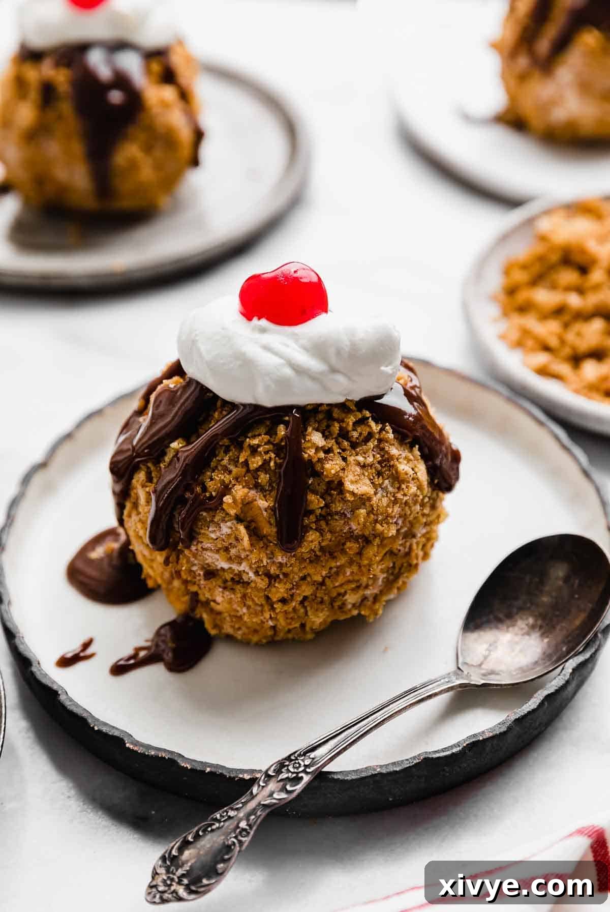 Golden Frost 6 Fried Ice Cream ball on a black rimmed white plate, the no fried fried ice cream is topped with hot fudge drizzle, whipped cream dollop, and a maraschino cherry.