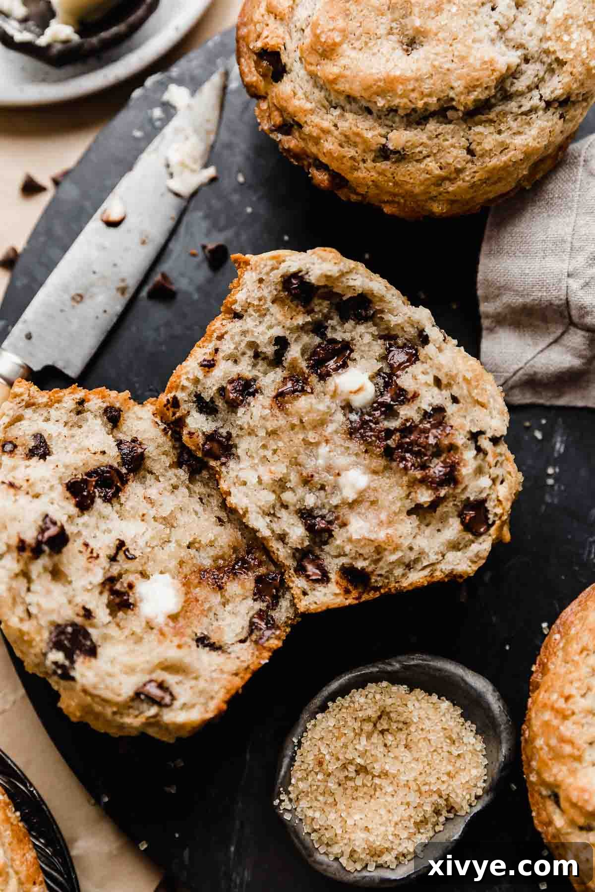 A Bakery Style Chocolate Chip Muffin cut in half and topped with softened butter, on a black background for a dramatic effect.