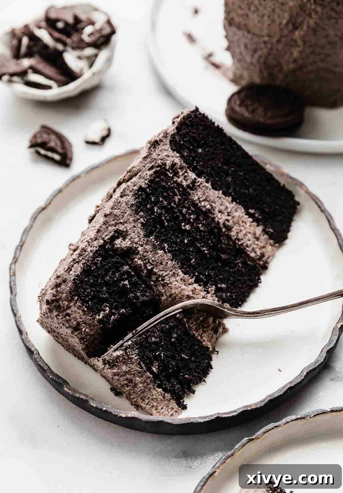 A slice of elegant three-tiered Oreo cake on a black-rimmed white plate, with a fork gently cutting into the top portion of the cake, highlighting its moist layers and creamy frosting.