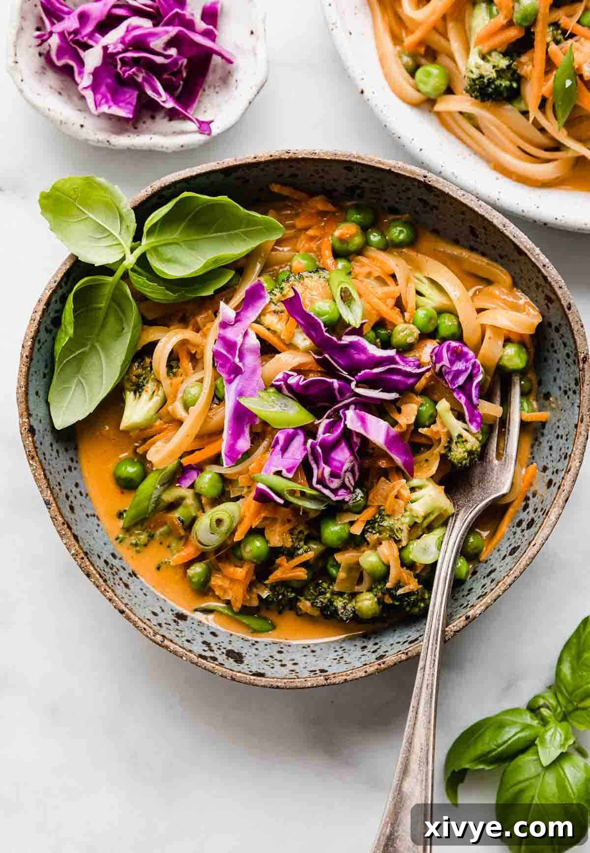 A blue bowl filled with vibrant Coconut Curry Noodles and an assortment of colorful vegetables, elegantly topped with fresh basil and finely shredded purple cabbage.
