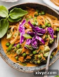 A blue bowl filled with red orange colored Coconut Curry Noodles with vegetables and topped with basil and purple cabbage.