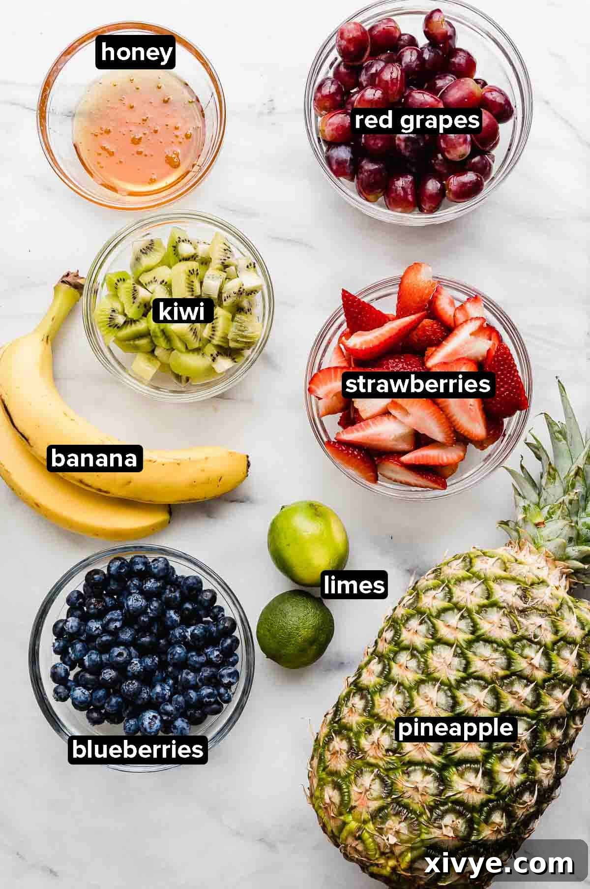 Vibrant Fruit Fusion 3 Fruit Cocktail ingredients displayed on a white background, including fresh pineapple, kiwi, strawberries, red grapes, limes, honey, bananas, and blueberries.