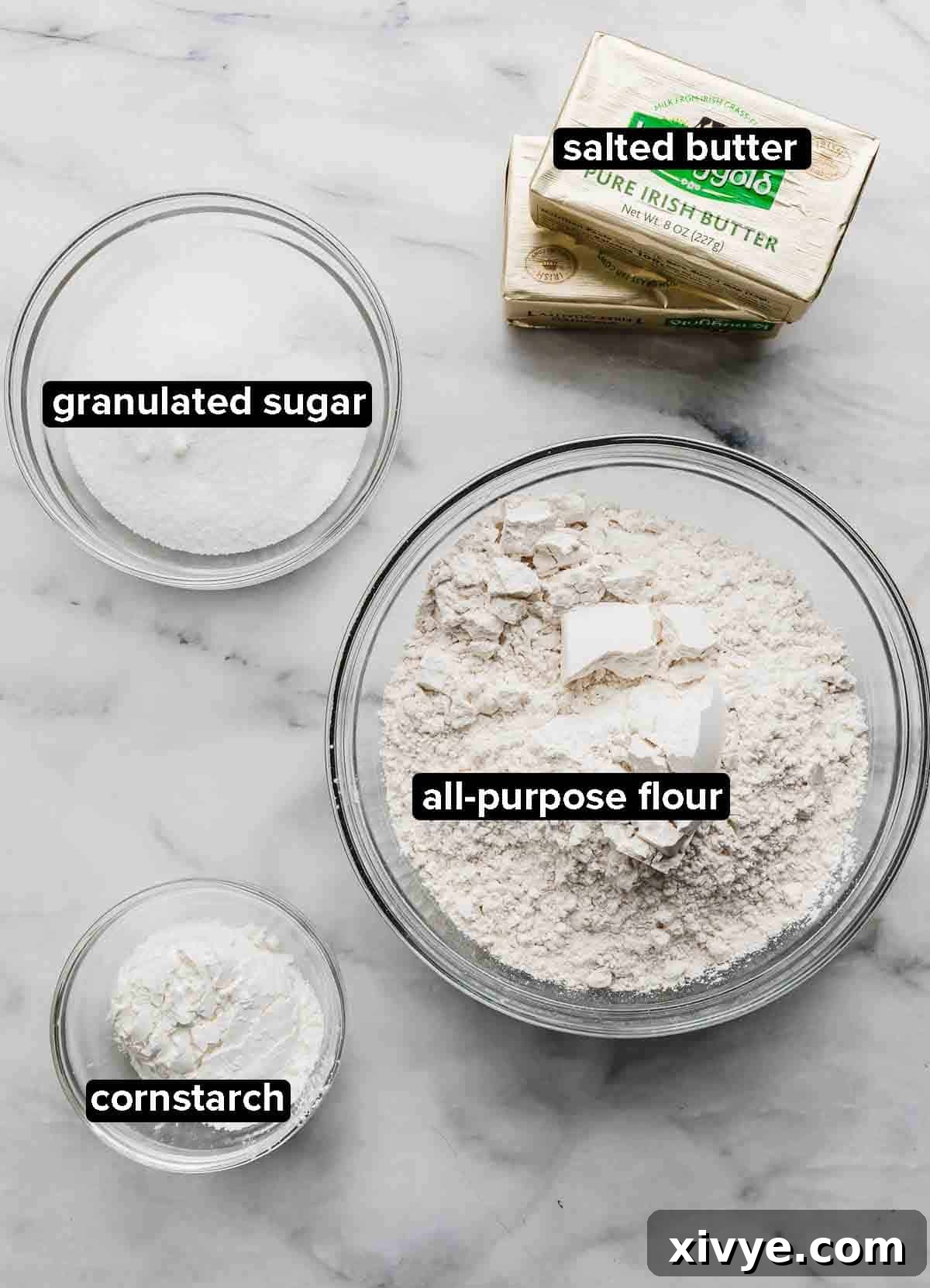 All the ingredients for Grandma's Shortbread Cookies, neatly portioned into glass bowls on a white and gray marble countertop: all-purpose flour, granulated sugar, cornstarch, and softened salted butter.