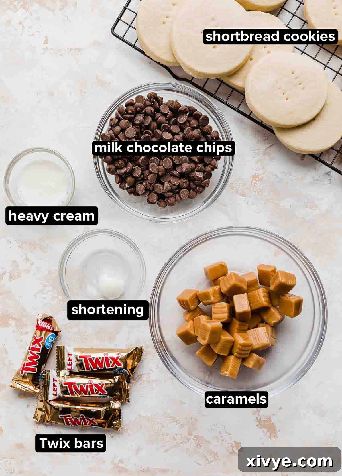 Twix Cookie ingredients on a white and cream textured background: shortbread cookies, caramels, chocolate chips, Twix bars, shortening, and heavy cream.