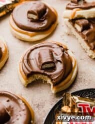 A Twix Cookies made with a shortbread cookie base, topped with caramel, chocolate and a Twix bar on a light brown background.