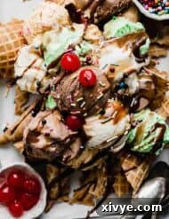 Scoops of chocolate, vanilla, and mint ice cream on top of waffle cones with hot fudge, and maraschino cherries on top.