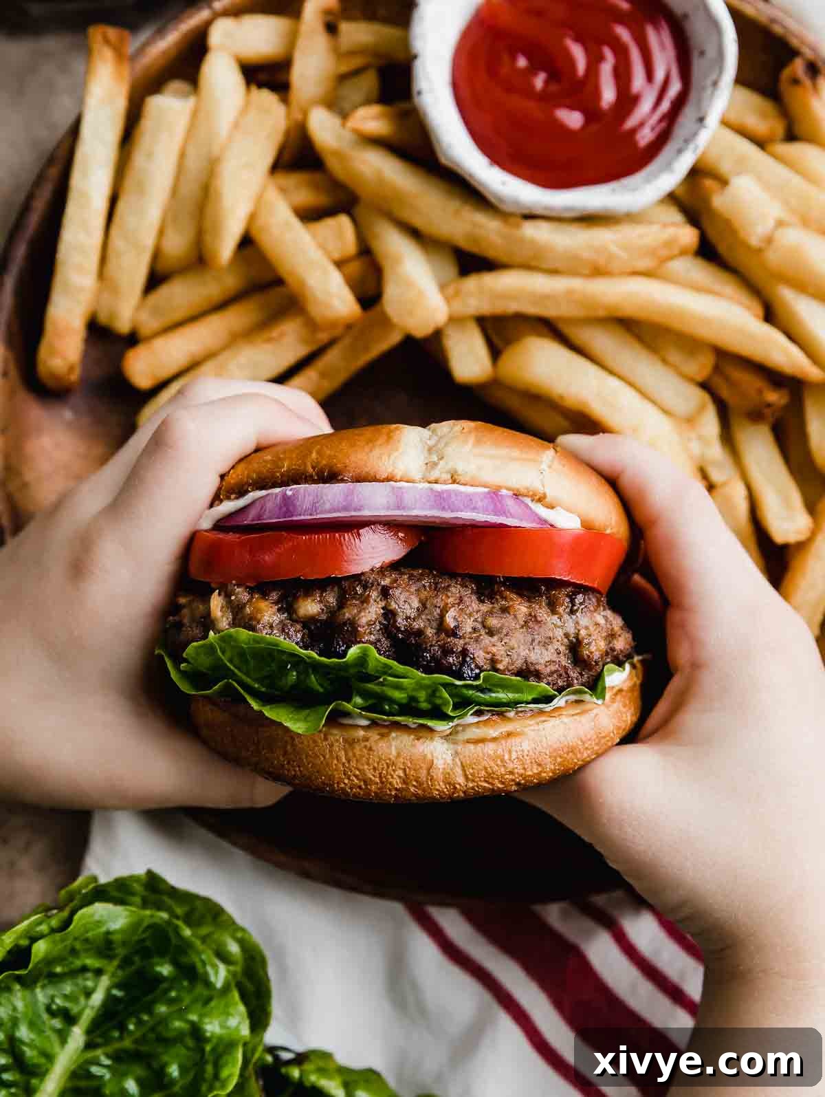Signature Steakhouse Burgers 6 Two hands holding a Steakhouse Burger on a bun with lettuce, tomato, and purple onion, with French fries in the background.