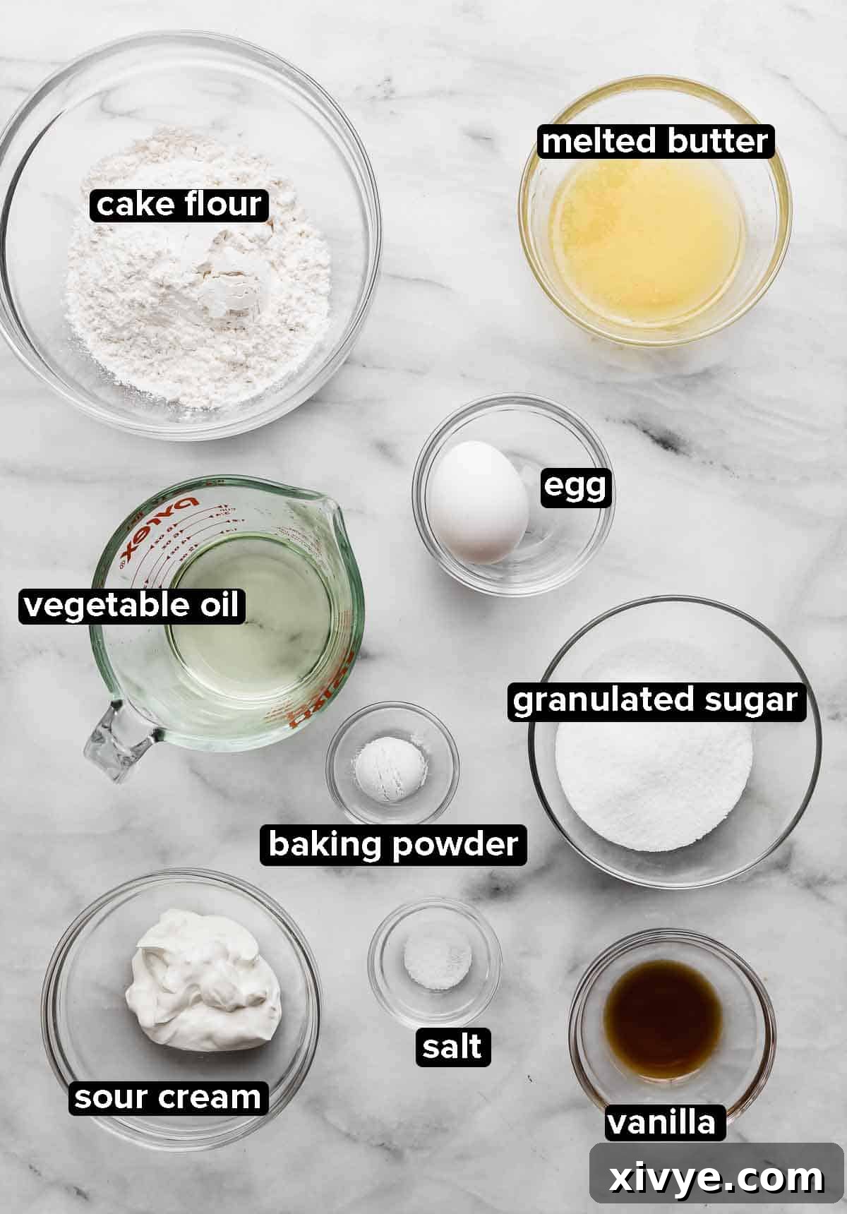 Bite-Sized Delights 3 Vanilla Mini Cupcakes ingredients portioned into glass bowls on a white marble background, ingredients include: vegetable oil, cake flour, butter, egg, sugar, sour cream, vanilla, salt, baking powder.
