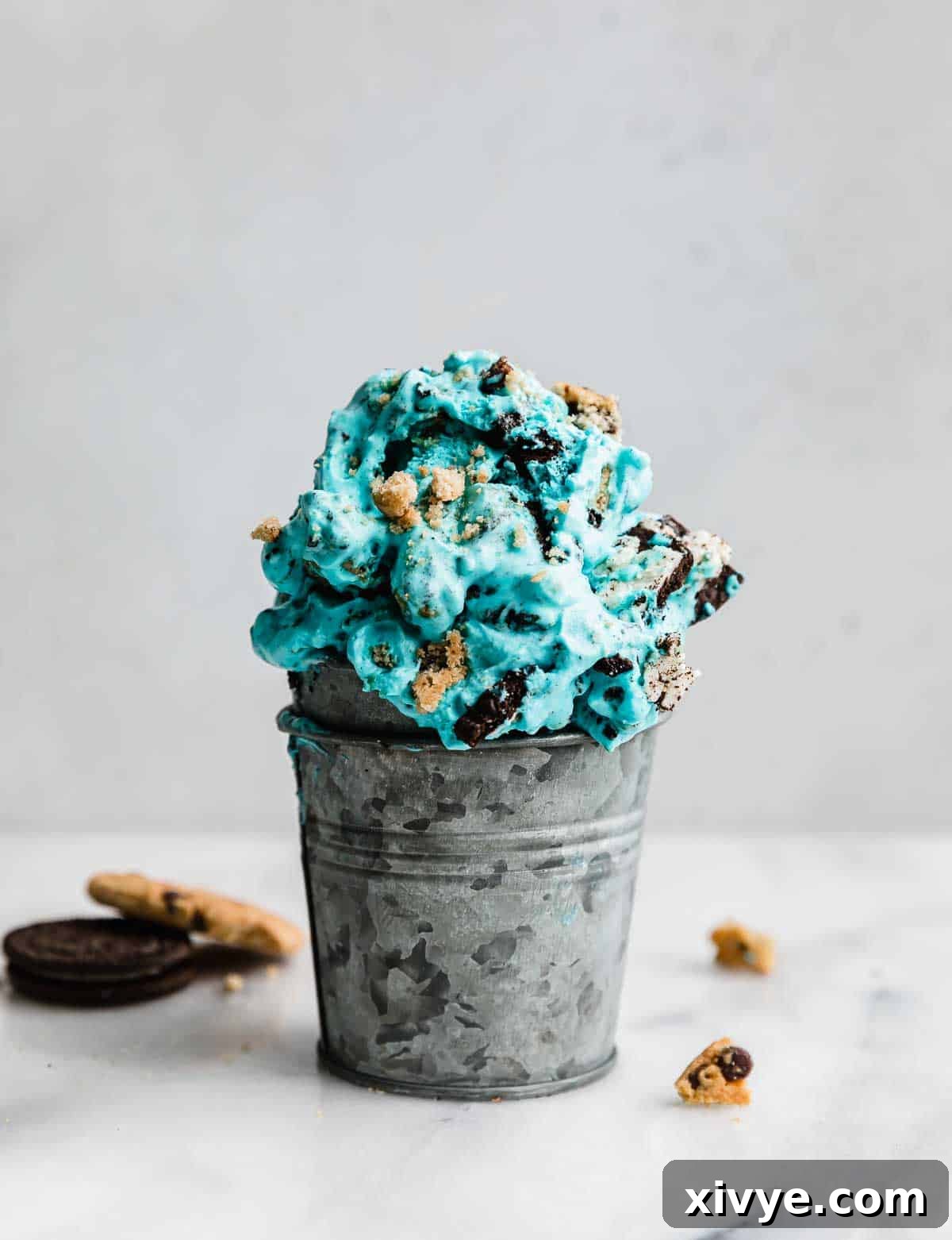 A small metal cup brimming with vibrant blue Cookie Monster Ice Cream, showcasing generous chunks of Oreo and chocolate chip cookies, set against a pristine white background. A perfect visual representation of this fun, homemade dessert.