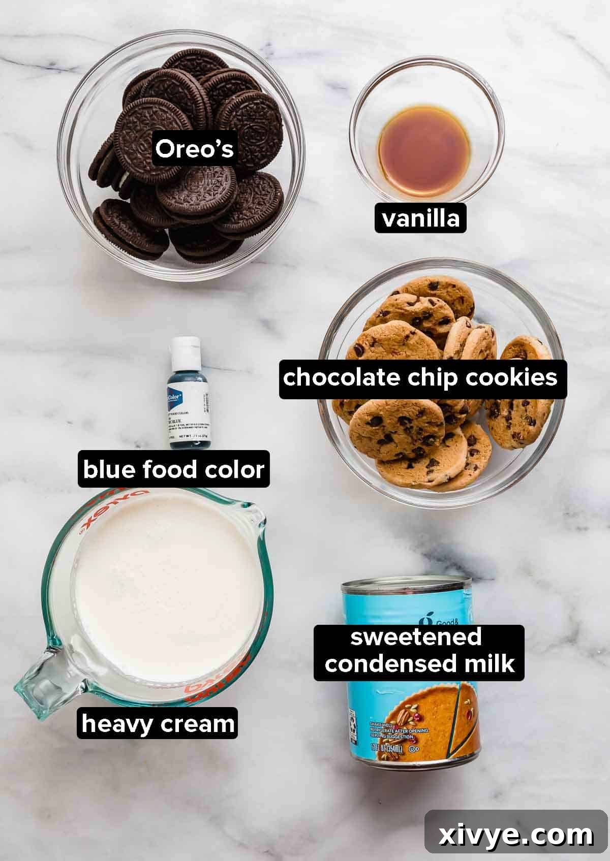 A beautifully arranged flat lay of ingredients on a white marble background, ready for making Cookie Monster Ice Cream. Featured items include classic Oreo cookies, crunchy chocolate chip cookies, pure vanilla extract, a carton of rich heavy cream, and a can of sweet condensed milk, highlighting the simplicity and quality of components.