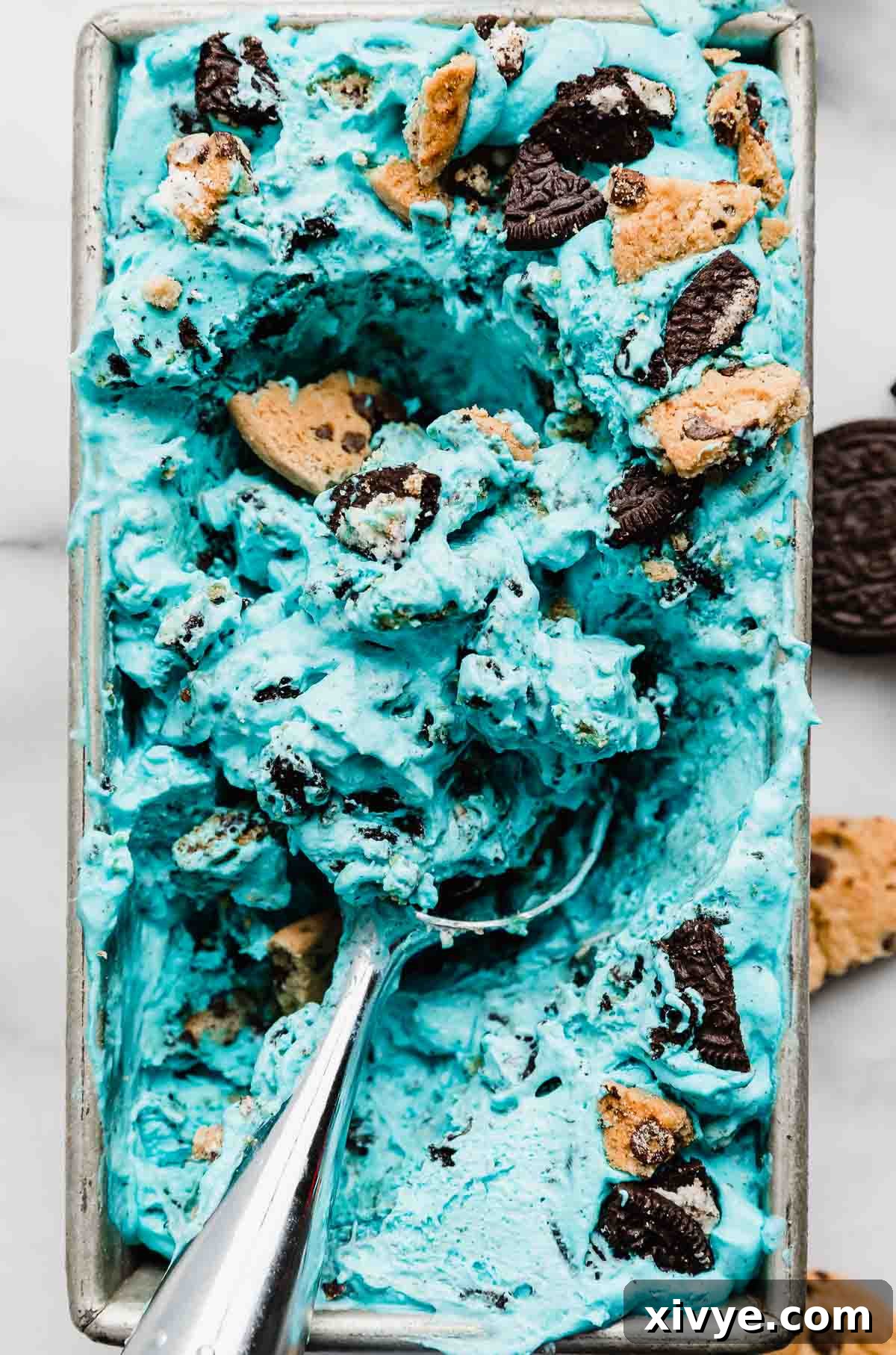 An ice cream scooper deftly carving into a pan of no-churn blue Cookie Monster Ice Cream, laden with visible chunks of cookies, ready to be served. The image captures the creamy texture and deliciousness of the homemade dessert.