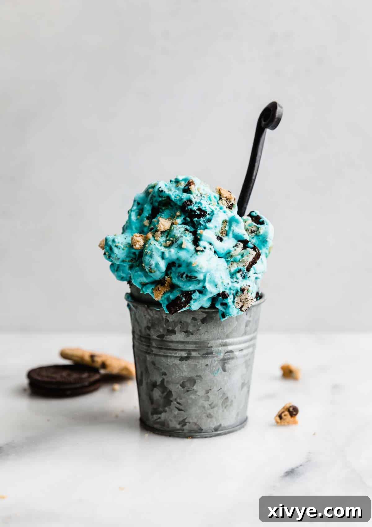 A charming metal cup filled to the brim with vibrant blue Cookie Monster Ice Cream, showcasing the enticing mix of cookie chunks. The cup is artfully placed against a clean, white background, emphasizing the dessert's appealing color and texture.