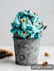 A close-up shot of a metal cup overflowing with Cookie Monster Ice Cream, generously studded with chunks of Oreo and chocolate chip cookies. The vibrant blue color and abundant cookie pieces make it an incredibly tempting treat.