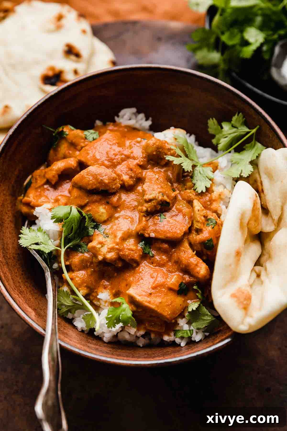 The Creamy Tikka Masterpiece 6 A serving of Chicken Tikka Masala with cilantro garnish and naan bread on a rustic background.