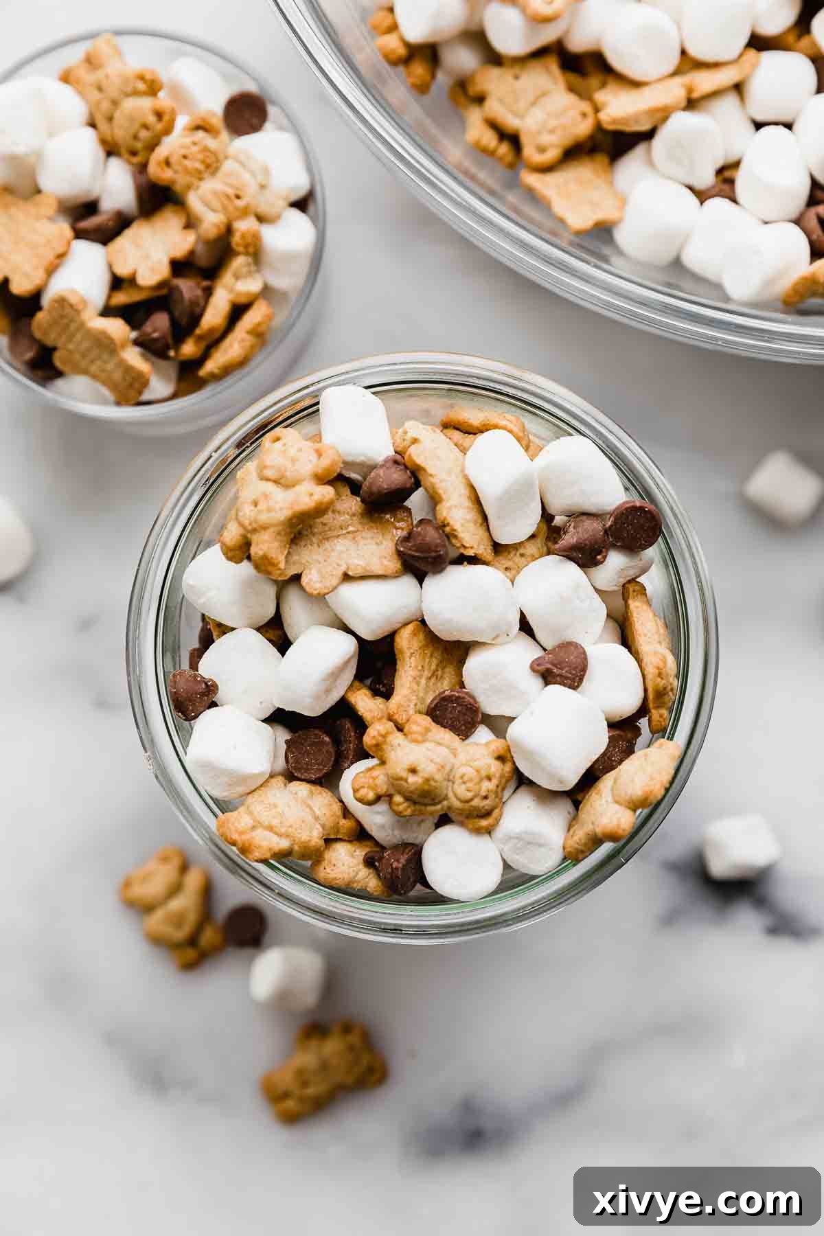 Smores Trail Mix 2 S'mores Mix in a clear glass cup on a pristine white marble background, showcasing a delectable combination of crunchy Teddy Grahams, soft mini marshmallows, and rich milk chocolate chips, ready to be enjoyed as a perfect summer snack.
