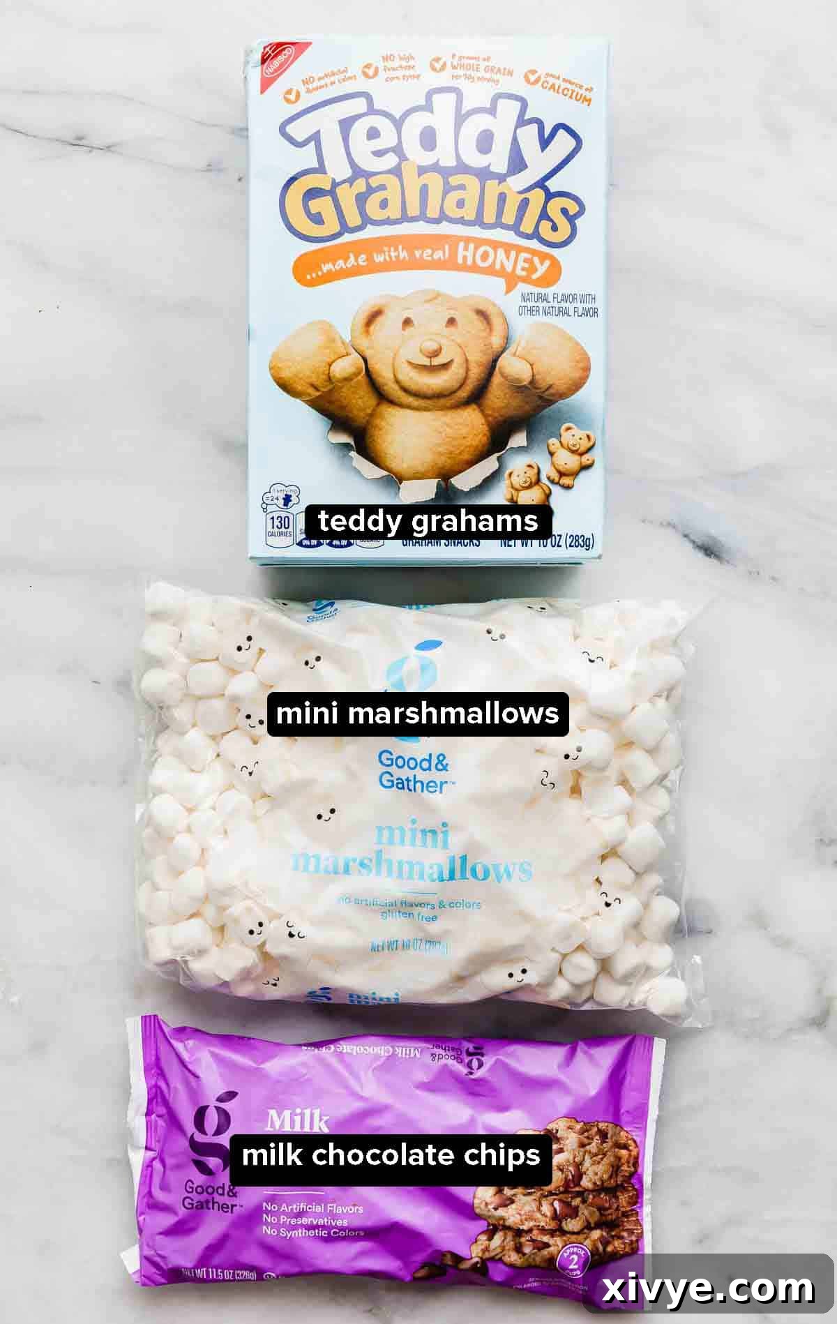 Smores Trail Mix 3 S'mores snack mix ingredients laid out on a white background, featuring a box of honey Teddy Grahams, a bag of mini marshmallows, and a bag of rich milk chocolate chips, ready for assembly.