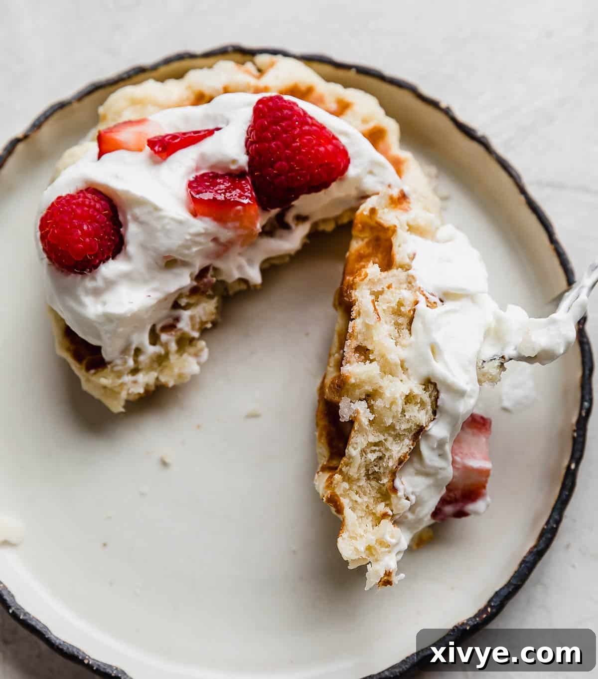 A Belgian Liege Waffle topped with whipped cream and chopped strawberries and raspberries, with a fork cutting into the center of the waffle, highlighting its delicious texture.