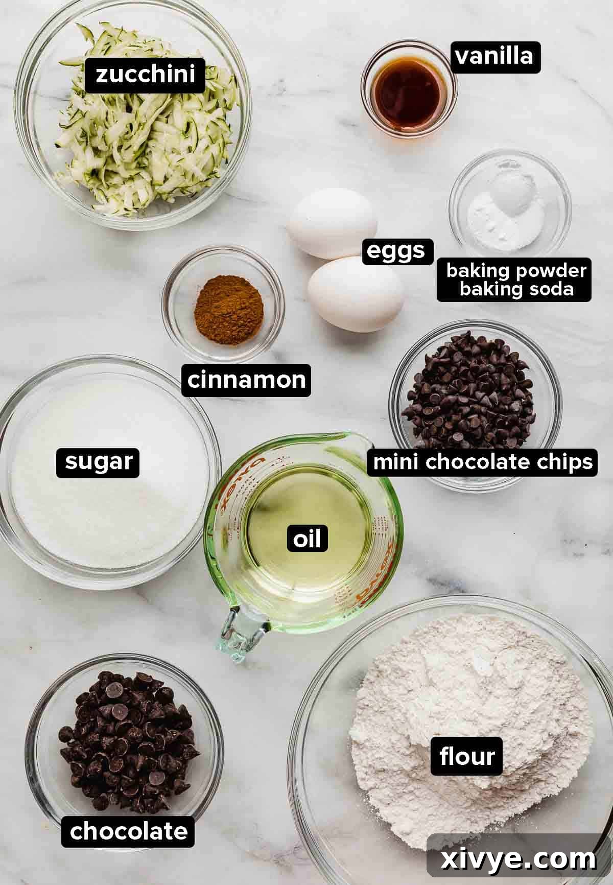 Garden Delight Chocolate Chip Muffins 3 Zucchini Chocolate Chip Muffins ingredients on a white and gray marble background: shredded zucchini, oil, egg, flour, chocolate chips, sugar, vanilla, cinnamon, and baking soda and baking powder.
