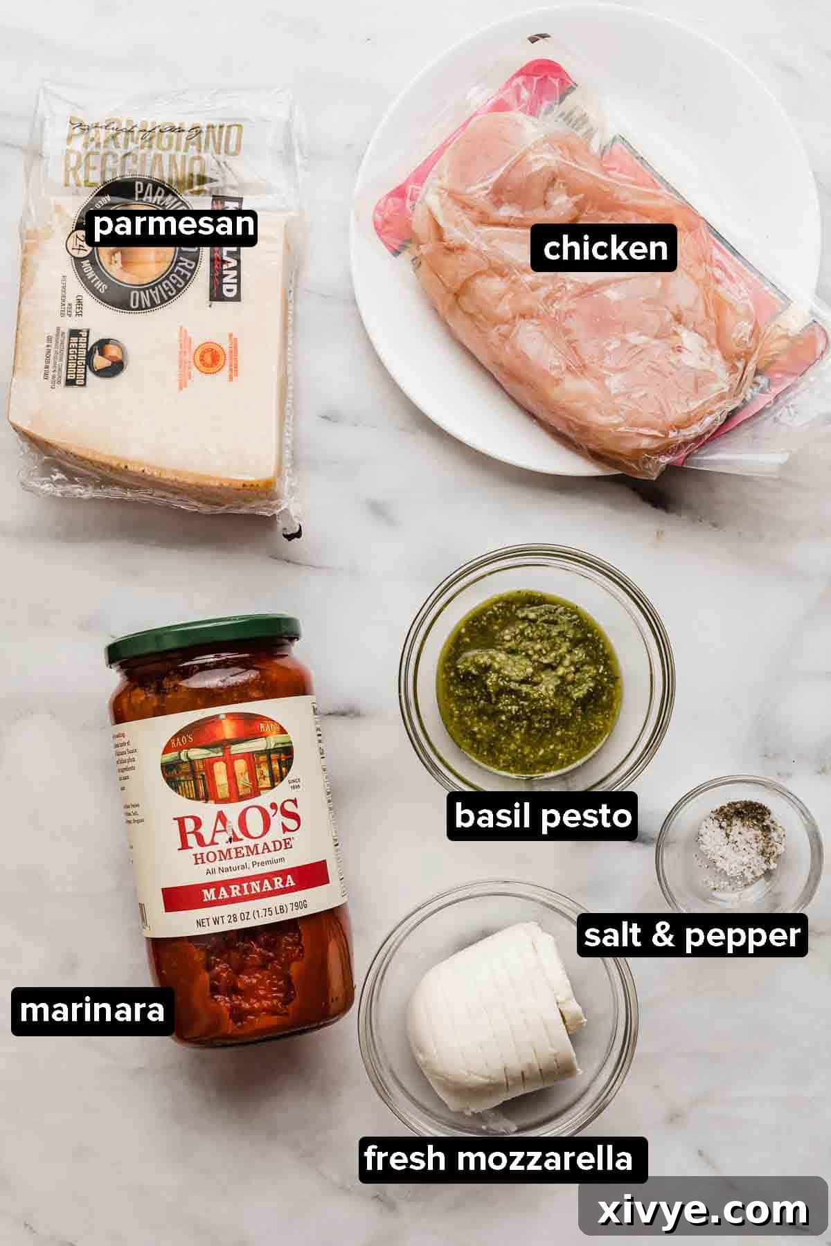Pesto Chicken Parmesan ingredients on a white marble table, parmesan, chicken, marinara, pesto, mozzarella, salt and pepper.
