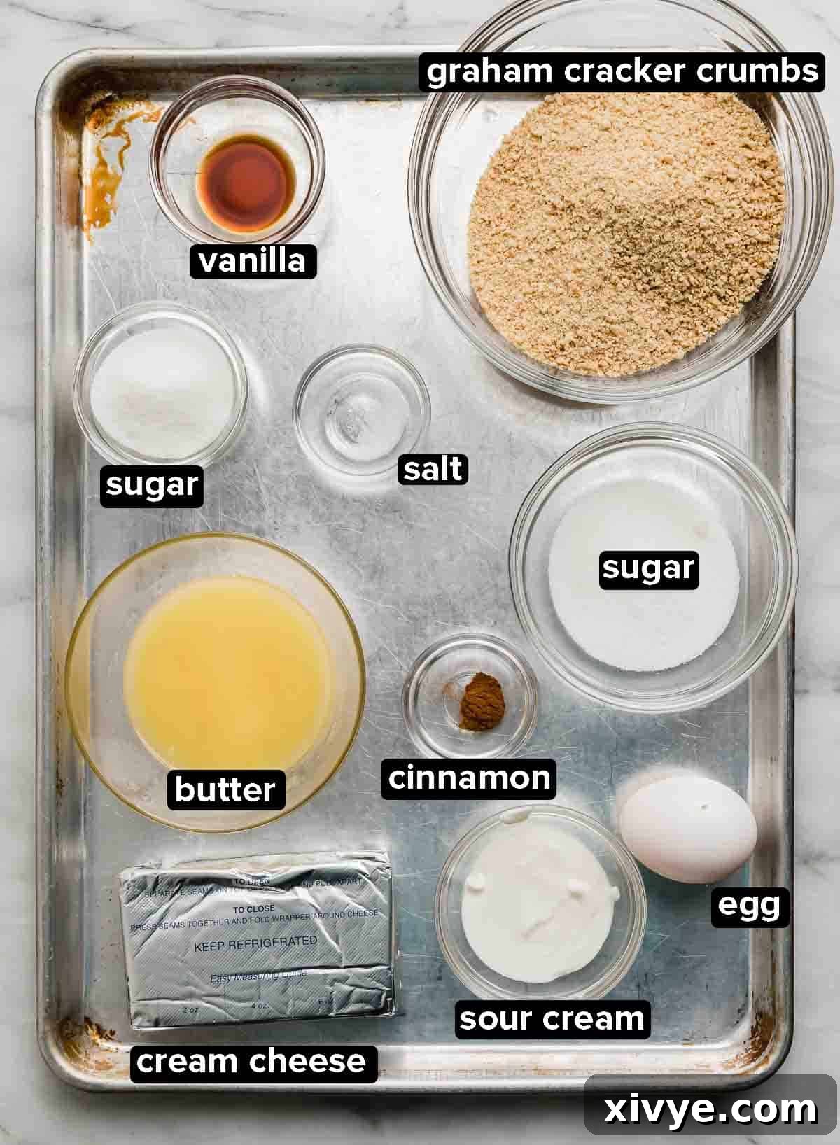 Ingredients used to make the best Mini Cheesecakes recipe portioned into glass bowls on a silver baking sheet: graham cracker crumbs, butter, sugar, egg, vanilla, and cream cheese.