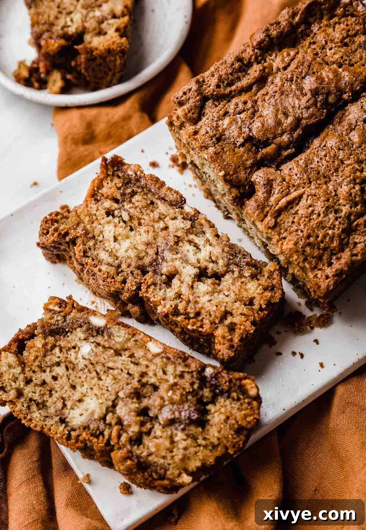 A loaf of freshly baked Cinnamon Swirl Bread with visible white chocolate chips and a golden-brown crunchy topping, resting on a pristine white rectangular plate.