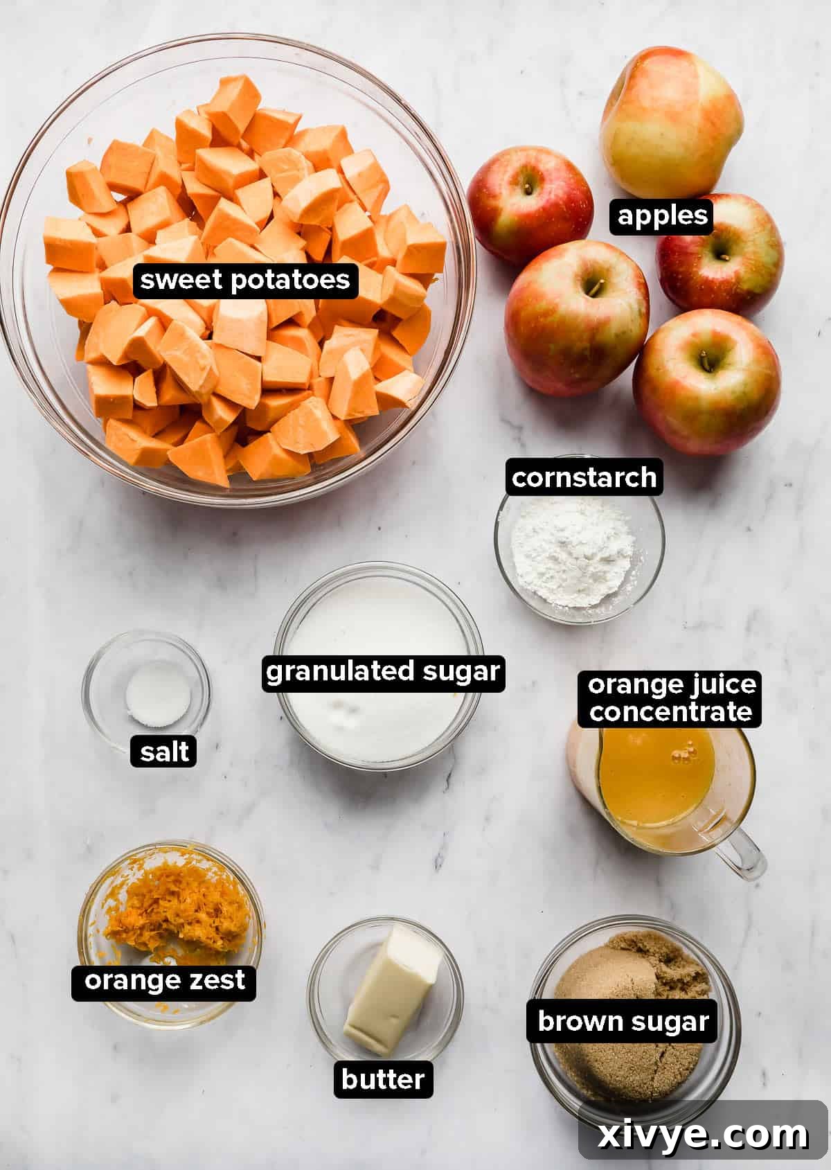 Assortment of candied yam ingredients in glass bowls, including sweet potatoes, apples, oranges, and spices, on a elegant marble background.
